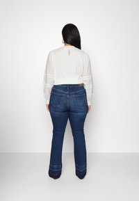 Blusa branca de manga longa com decote franzido e detalhe em forma de buraco, combinada com jeans azuis boca de sino com bainha costurada em contraste e bolsos.