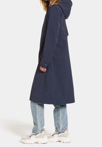 Manteau long imperméable bleu marine avec capuche, présentant une coupe à épaules tombantes, des poignets à boutons et des poches lat érales. Associé à un jean clair.