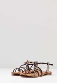 Sandali neri con punta aperta e cinturini incrociati, con un design in rete testurizzata e una soletta in pelle liscia. Tacco in legno marrone chiaro.