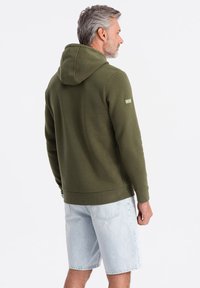 Sudadera verde oliva hecha de un tejido suave con un bolsillo canguro. Presenta una capucha grande y un dobladillo acanalado, combinada con pantalones cortos de mezclilla claros.