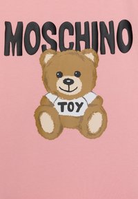 Růžová látka s kresleným medvědem hnědé barvy a černýma očima, který má na sobě bílou košili s nápisem "TOY" a vedle ní tučným černým textem "MOSCHINO."