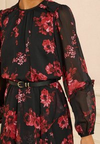 Rochie neagră cu flori, având flori roșii și roz, cu un guler franjurat, mâneci lungi și o centură neagră în talie.