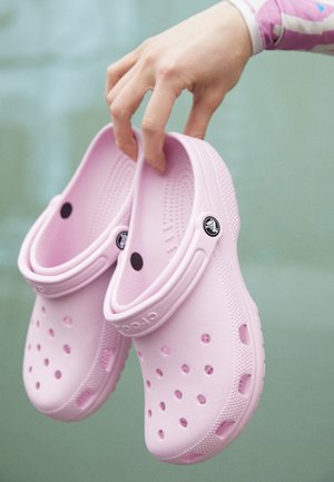 Main tenant une paire de sabots Crocs rose clair avec des trous de ventilation et des brides de talon réglables sur un fond vert flou.