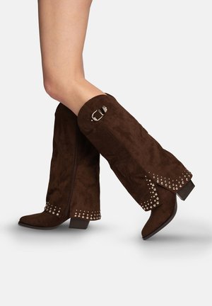 Benen met kniehoge, donkerbruine suède laarzen met zilveren studs en een decoratieve gesp, getoond in een looppositie op een witte achtergrond.