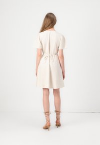 Femme debout de dos face à la caméra, vêtue d'une robe crème à manches courtes avec une ceinture à la taille et des sandales à talons beige lacées. Fond uni.