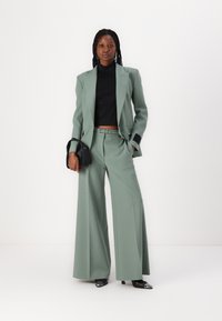 Zöld, testre szabott öltöny, széles lábú nadrággal és kétsoros blézerrel. Fekete crop top alatta. A modell egy fekete táska-tart.
