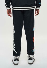 Schwarze Baumwoll-Sweatpants mit weißer Schrift und bunten Grafiken. Mit elastischen Bündchen und seitlichen Streifen am Bund.