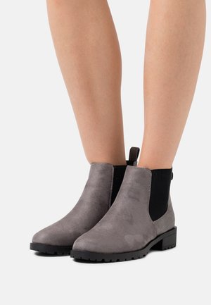 Botas de tobillo de ante gris con paneles laterales de elastano negro y suelas dentadas, usadas con piernas al descubierto, fotografiadas sobre fondo blanco.