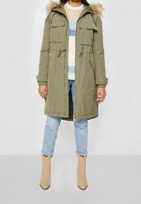 Olivově zelená parka s kapucí s umělým kožíškem, dvěma náprsnými kapsami, stahovacím pasem a zapínáním na zip, nošená přes pruhovaný svetr a světlé džíny.