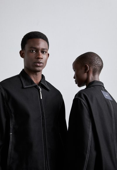 Schwarze Zip-Jacke aus dickem Material mit weißen Stichdetails. Verfügt über einen Kragen und ein Etikett auf der Rückseite, dargestellt an zwei Figuren.