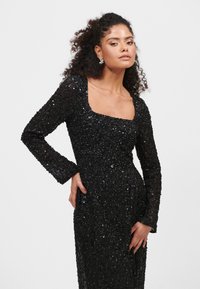 Abito lungo nero a maniche lunghe con scollo quadrato. Vestibilità aderente, superficie testurizzata e silhouette elegante. Accenti scintillanti notevoli in tutto l'abito.