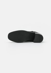 Suela de zapato negra con una superficie texturizada, punta redondeada y un tacón plano. Presenta un acabado suave y contornos de diseño sutiles.