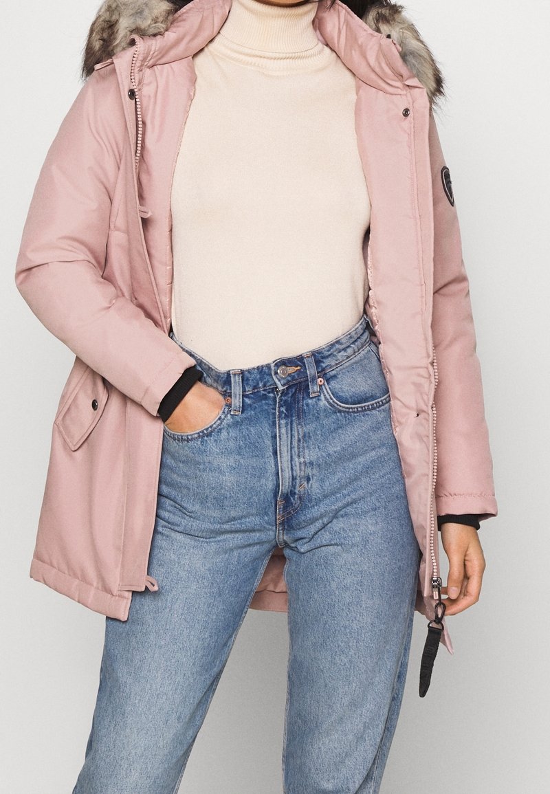 Rosa vadderad parka med en foderkrage av päls, dragkedja och knappstängning samt sidofickor. Bärs över en beige polotröja och blå högmidjade jeans.