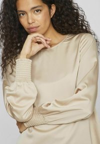 Beige satinblus med rund halsringning, puffärmar och smockade manschetter. Slät textur med mjuk drapering och minimalistisk design.