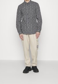 Camicia a righe bianche e nere con bottoni e maniche lunghe, abbinata a pantaloni cargo beige con tasche e una toppa con logo, indossata con scarpe da ginnastica nere.