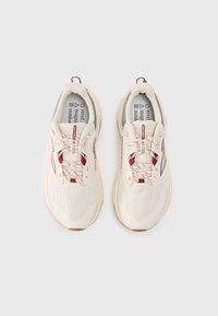 Chaussures de sport beige clair avec une tige en maille respirante, des accents rouges, un système de laçage et une semelle en caoutchouc texturée. Affiche une étiquette de marque bien visible.