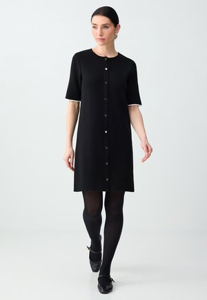 Femme marchant vêtue d'une robe noire boutonnée à manches courtes, de collants noirs et de chaussures Mary Jane noires, sur un fond clair uni.