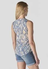 Mujer con cabello largo y rubio, lleva grandes pendientes de aro, una blusa sin mangas con estampado y shorts de mezclilla remangados, vista desde atrás.
