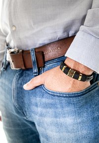 Black and gold bracelet made of woven material, worn on a wrist, with a gold-accented clasp, paired with a blue denim shirt and brown belt.