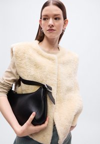 Μπεζ γιλέκο από shearling με στρογγυλεμένες άκρες, φορεμένο πάνω από ένα ελαφρύ πουλόβερ. Το μοντέλο κρατάει μια μαύρη δερμάτινη τσάντα με καμπυλωτό σχήμα και ασημένια μεταλλικά μέρη.