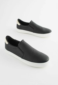 Chaussures noires à enfiler avec une surface texturée, semelles en caoutchouc blanches et accents métalliques dorés au talon. Sans lacets, design minimaliste.