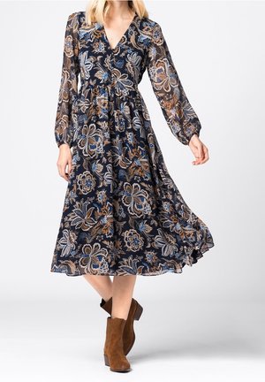 Femme portant une robe midi bleu marine à fleurs avec des manches longues en voile transparent et des bottines marron, debout devant un fond uni.