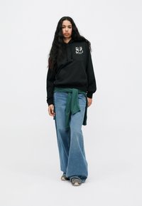 Fekete kapucnis pulóver grafikával, világoskék, bő szárú farmernadrág, zöld ing a derekán megkötve, és szürke sportcipő. Általánosan sima textúra.