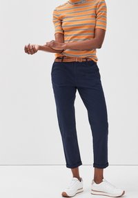 Chemise à rayures orange et blanches à manches courtes, pantalon bleu marine et baskets blanches avec des accents beige. La chemise a une texture côtelée et une coupe slim.