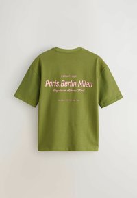 Olíva zöld póló rózsaszín felirattal, amelyen a "Párizs. Berlin. Milanó" és a "Euphoria Waves Fest" szöveg látható a hátsó részén, népszerűsítve egy zenei fesztivált.