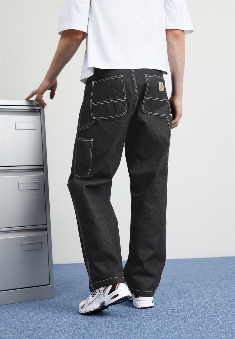 Pantalones de mezclilla negros con costuras blancas en contraste, diseño de pierna recta, múltiples bolsillos y una textura suave, combinados con zapatillas blancas.