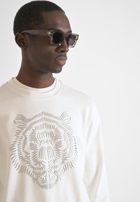 Gráfico de tigre gris en una sudadera blanca; el modelo lleva gafas de sol oscuras y angulares. Tejido suave con un cuello alto.
