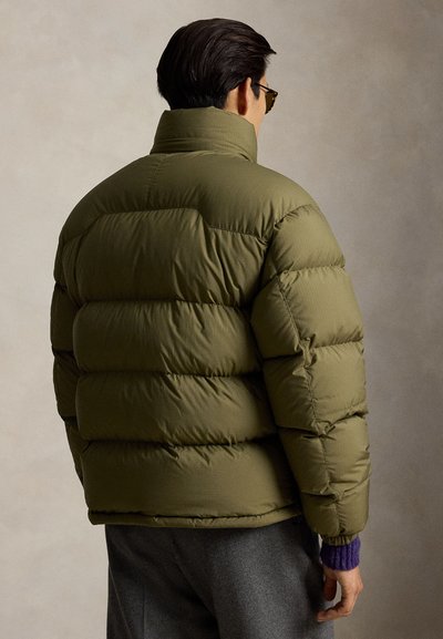 Homme portant une doudoune vert olive et un pantalon gris foncé, debout de dos sur un fond neutre.