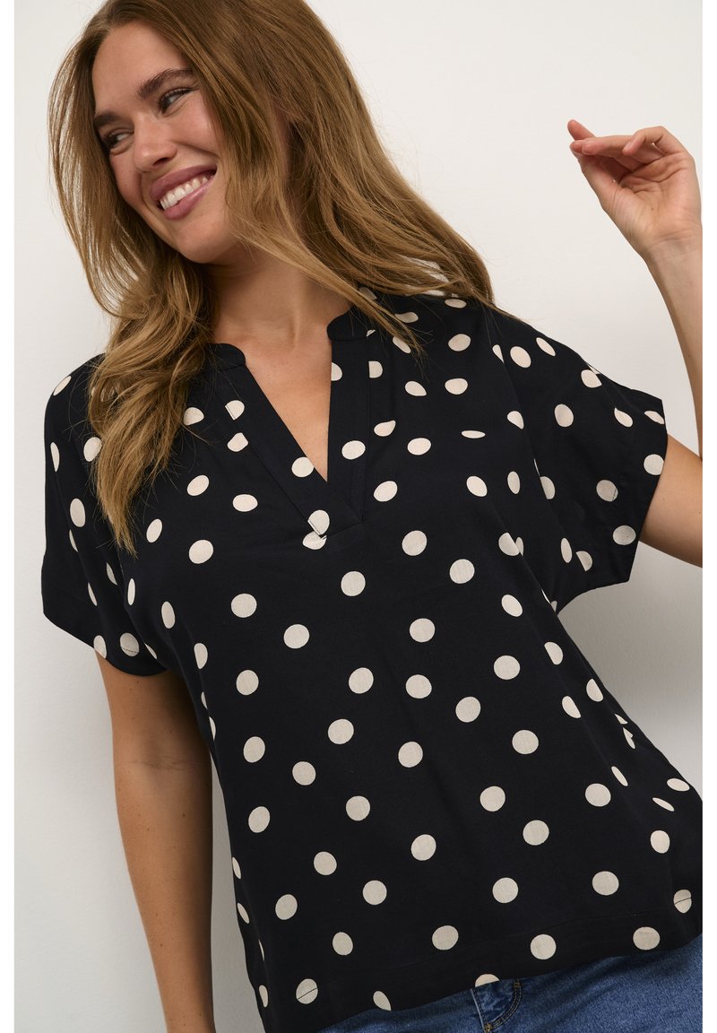 Blouse noire à manches courtes avec un col en V, présentant un motif à pois blancs et une coupe décontractée. Tissu lisse avec un léger brillant.