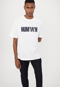 White cotton T-shirt featuring a bold black "HUMAN" logo, short sleeves, and a relaxed fit, paired with dark blue denim pants.