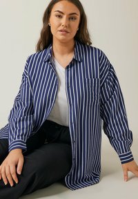 Camisa de botones a rayas navy y blanca con un bolsillo en el pecho, corte relajado y mangas largas, combinada con una camiseta blanca y pantalones negros.