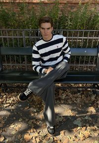 Jonge man in zwart-wit gestreepte trui en grijze broek zit op de handpalm in kleermakerszit omgeven door gevallen herfstbladeren.