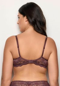 Donna con lunghi capelli scuri che indossa un reggiseno in pizzo viola e una mutandina abbinata, mostrata di spalle su uno sfondo semplice.