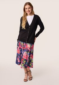 Zwarte gebreide cardigan met v-hals, knoopsluiting, gecombineerd met een fleurige, knielange rok in levendige kleuren en zwarte sandalen met bandjes.