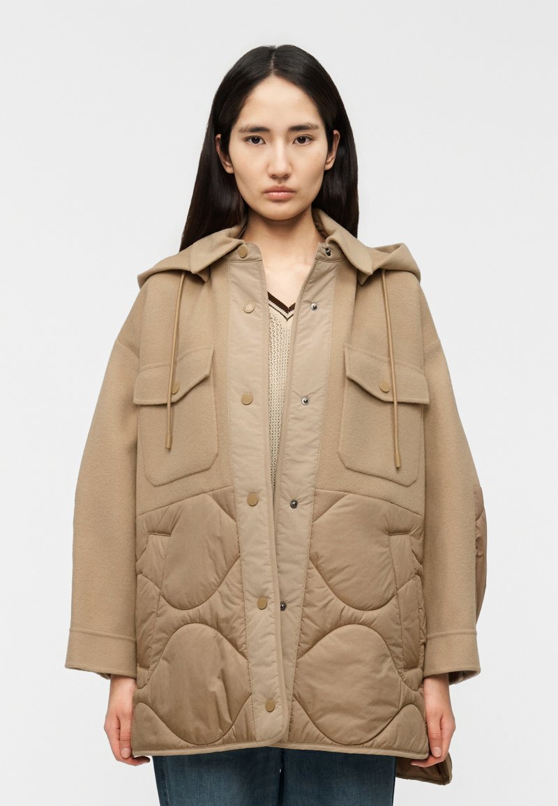 Μπεζ παλτό με καπιτονέ σχέδιο, κουκούλα, κούμπωμα με κουμπιά και δύο μπροστινές τσέπες. Απαλή υφή, άνετη εφαρμογή και πολυεπίπεδο σχέδιο με αντίθετο πάνελ.