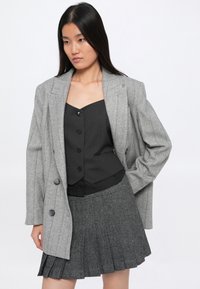 Femme portant un blazer gris à rayures sur un gilet noir à boutons et une jupe plissée grise, se tenant devant un fond blanc uni.