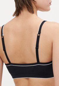 Juoda bralette su balta apdaila, pagaminta iš minkštos medžiagos. turi reguliuojamas petnešas ir paprastą, prigludusį dizainą. Be paminkštinimo ar lankelių.