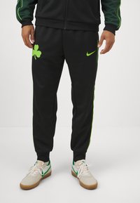 Fekete Nike melegítőnadrág zöld kiegészítőkkel és egy nagy zöld lóherével a bal lábon; keskeny fazonú és elasztikus mandzsettával készült.