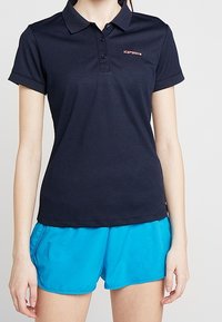 Mörkblå pikétröja i bomullsblandning, med en treknapplösning och en liten röd logotyp. Kombinerad med blå träningsshorts.