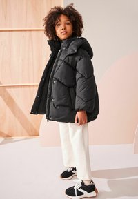 Schwarze Daunenjacke mit Kapuze, gesteppter Design und Druckknöpfen, kombiniert mit weißen Hosen und schwarzen Sneakers mit hellen Akzenten.