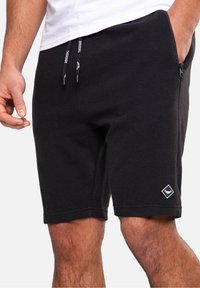 Homme portant un short en tricot côtelé noir avec taille à cordon et poche latérale zippée, main dans la poche, logo visible près de l'ourlet.