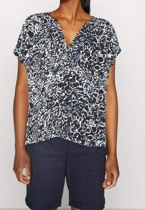 Frau trägt eine kurzärmelige Bluse mit schwarz-weißem abstraktem Muster und marineblaue knielange Shorts.