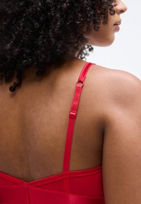 Κόκκινο σουτιέν με ρυθμιζόμενους ιμάντες, λεία υφή και δομημένο σχέδιο. Εμφανίζει την πίσω όψη και τις λεπτομέρειες στους ώμους.