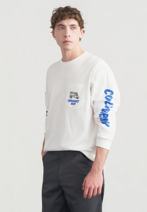 Άσπρο μακρυμάνικο T-shirt με τσέπη στο στήθος που διαθέτει γραφικό σχέδιο και μπλε κείμενο στα μανίκια· συνδυασμένο με σκούρο γκρι παντελόνια.