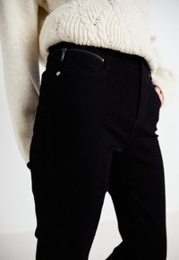 Jeans negros de tela texturizada, con bolsillo con cremallera y silueta ajustada. Combinados con un suéter de punto grueso color crema.