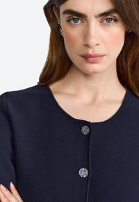 Cárdigan de punto azul marino con cuello redondo, presenta dos botones decorativos plateados en la parte frontal. Textura suave, diseño ajustado.